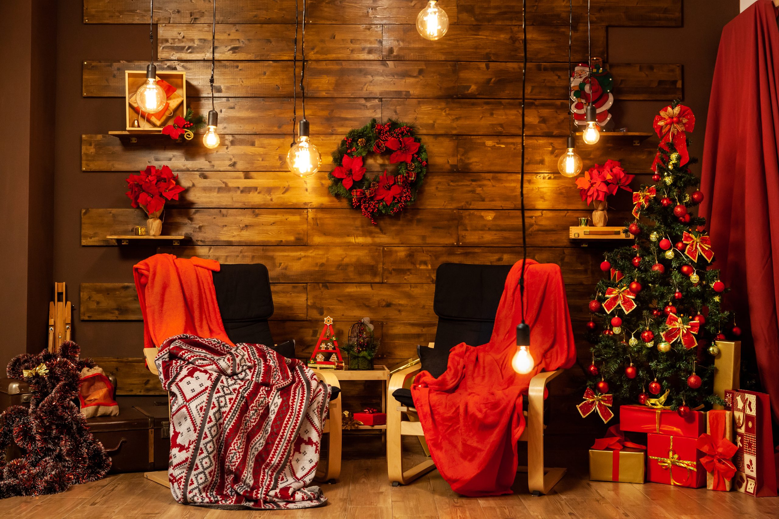 Decoração de Natal Rústica e Aconchegante com árvore clássica verde, guirlanda vermelha, luzes Edison pendentes e poltronas cobertas com mantas xadrez e vermelhas.