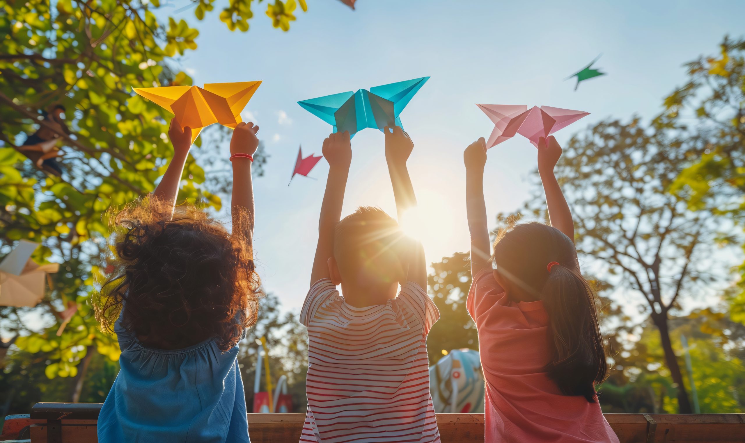 Três crianças brincando ao ar livre, levantando aviões de papel coloridos em direção ao céu, com o sol brilhando ao fundo e árvores ao redor, transmitindo alegria e liberdade.