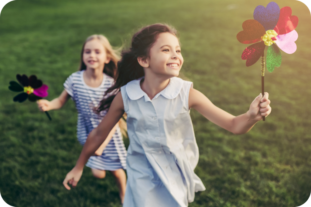 Duas meninas sorrindo correm em um gramado sob a luz do sol, segurando cata-ventos em formato de flor e expressando alegria, representando brincadeiras ao ar livre durante as férias.