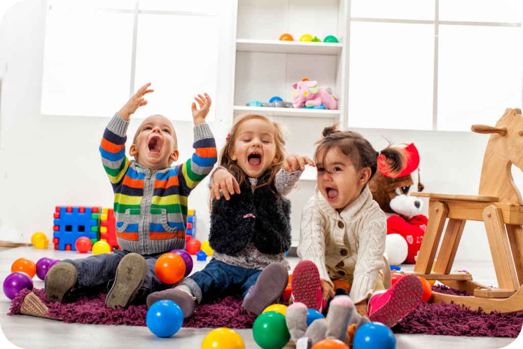 Três crianças pequenas (duas meninas e um menino) sentadas em um tapete cercadas por brinquedos e bolas coloridas em um ambiente indoor (fechado), gritando e rindo com entusiasmo, sugerindo diversão e brincadeiras em casa ou em espaços de lazer interno.